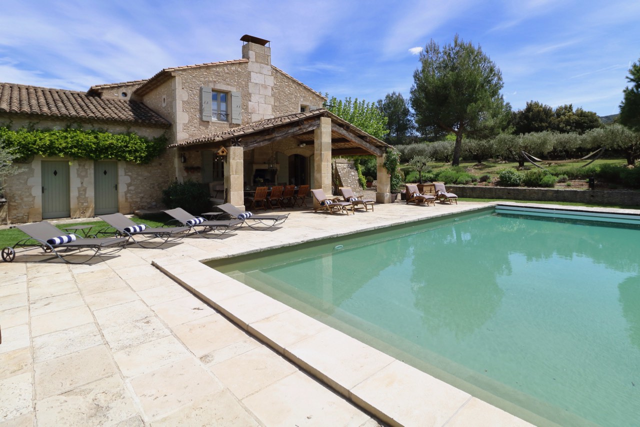 A traditional stone house with a blue swimming pool surrounded by sun loungers, set against a backdrop of lush green trees under a clear sky.